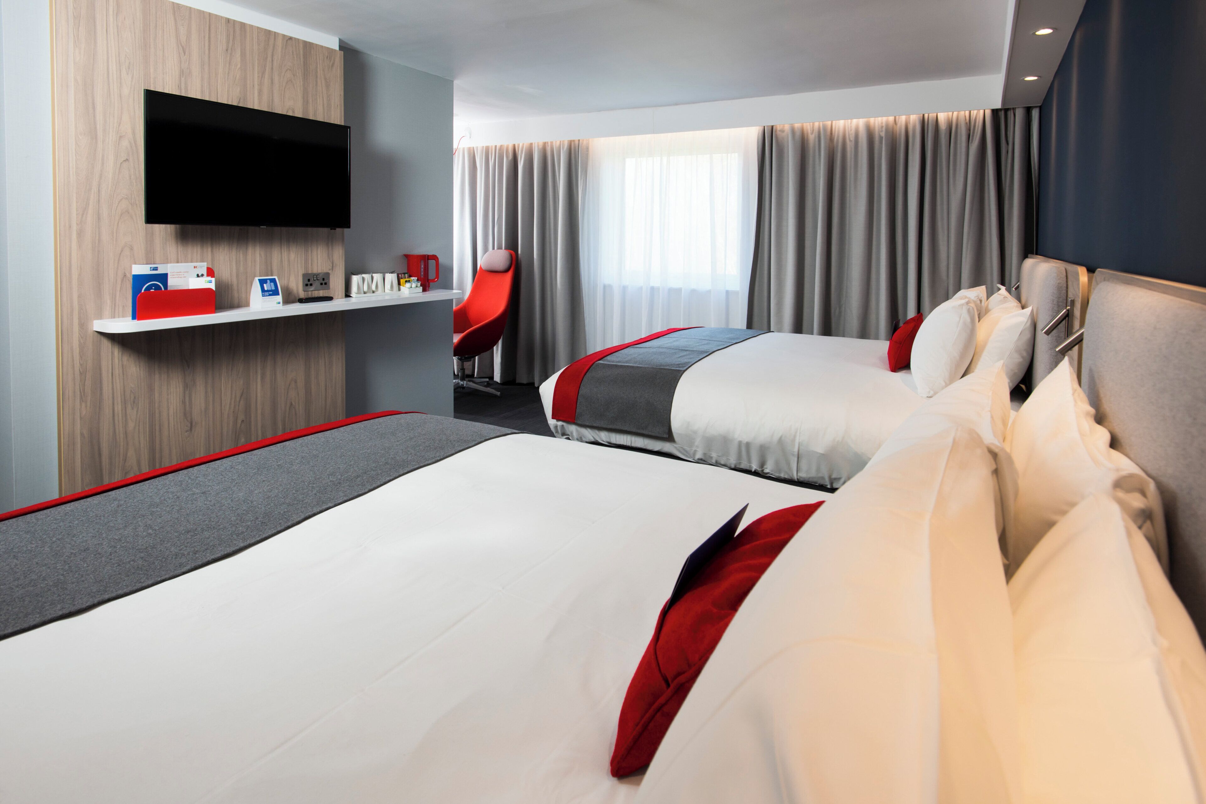 A hotel room with two beds, a flat-screen TV mounted on the wall, a small desk with a coffee maker, and a red chair.