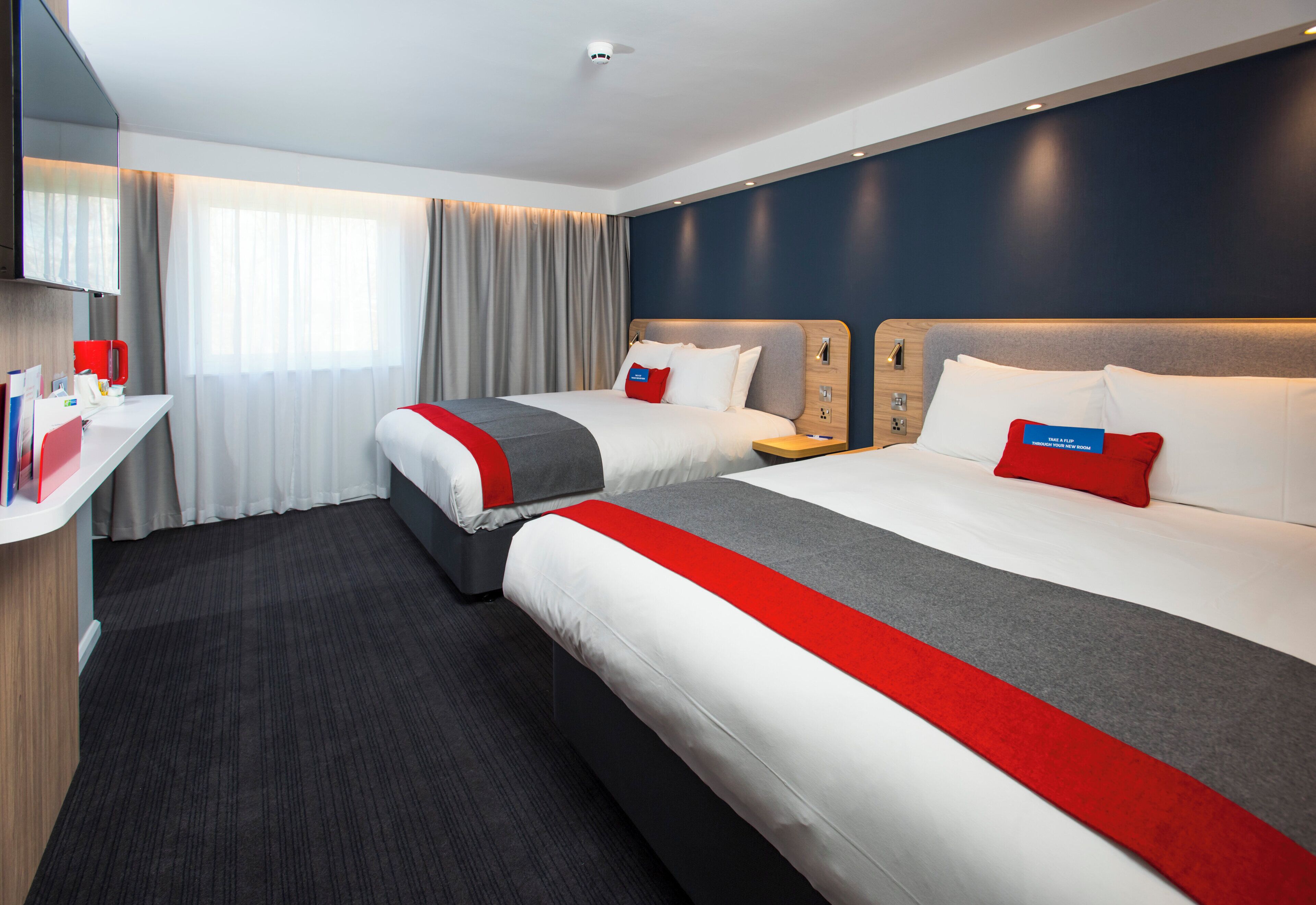 Habitació d'hotel amb dos llits, un escriptori, una televisió i una finestra amb cortines.