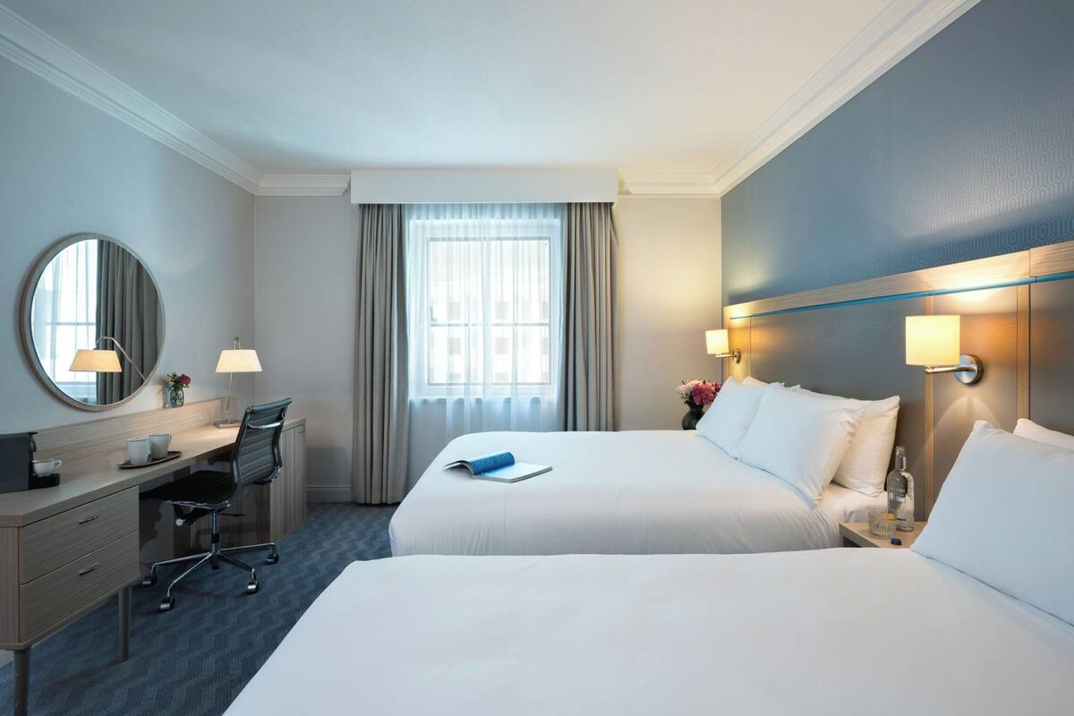 A hotel room with two beds, a desk, a chair, a round mirror, and a window with curtains.