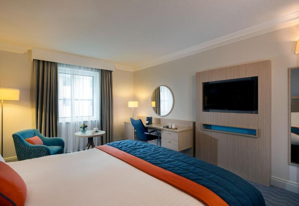 A hotel room with a large bed, a desk, a chair, a TV, and a window with curtains.