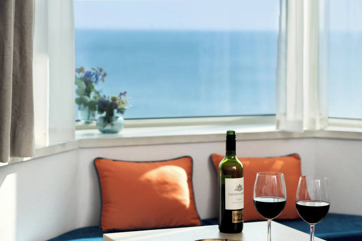 A table with a bottle of wine and two glasses of red wine, a window with a view of the sea, and a blue sofa with orange cushions.