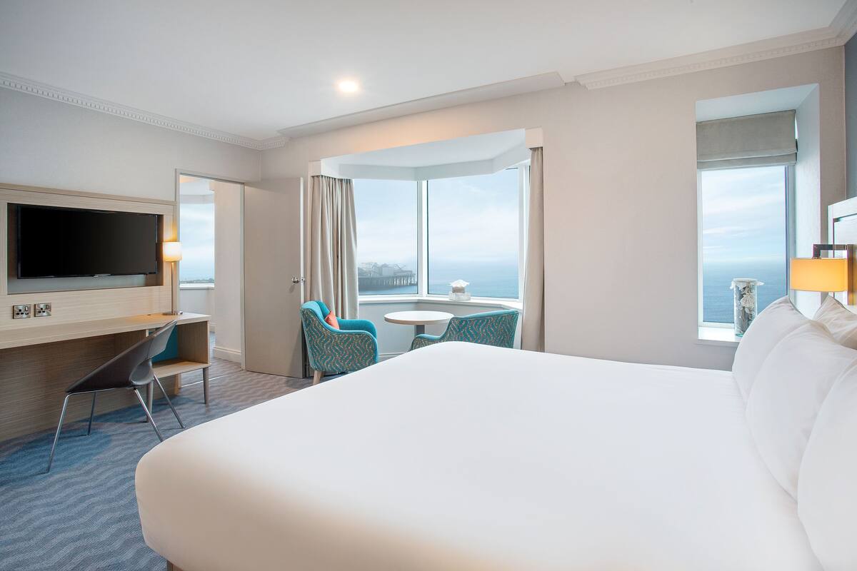 A hotel room with a large bed, a TV mounted on the wall, a desk with a chair, a small round table, and a view of the ocean through large windows.