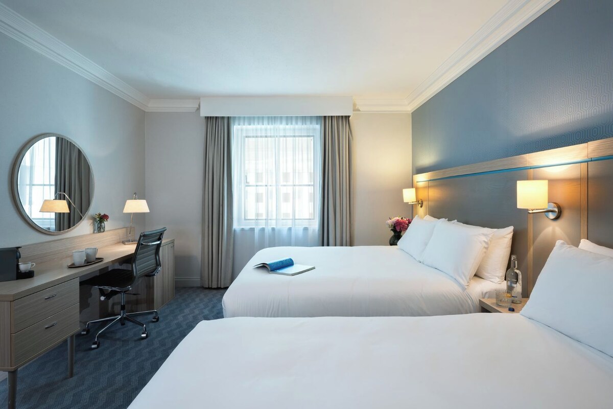 A hotel room with two beds, a desk, a chair, a mirror, and a window with curtains.