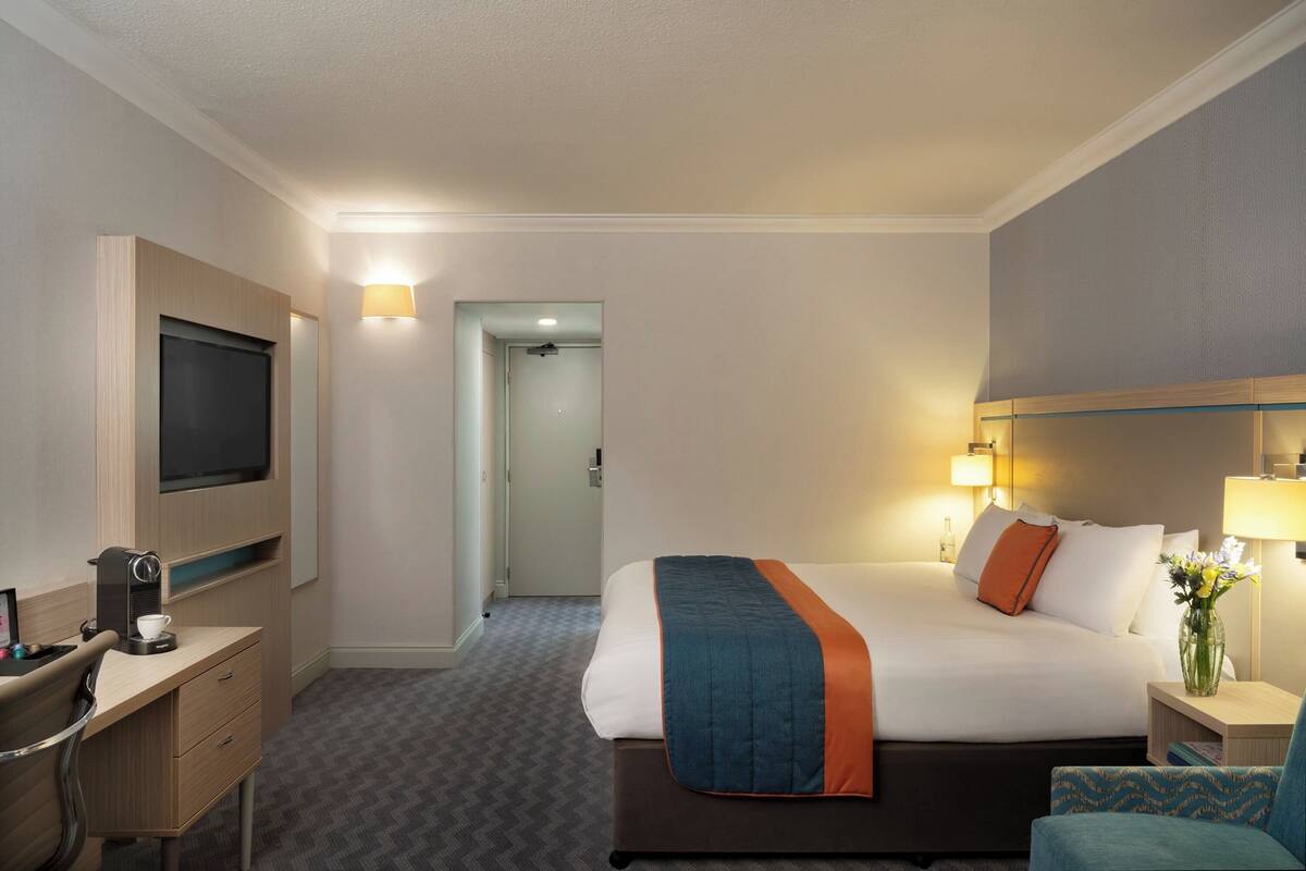 A hotel room with a bed, bedside table, lamp, TV, desk, chair, and a door leading to another room.