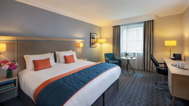 A hotel room with a large bed, a desk, a chair, a small table, and a window with curtains.