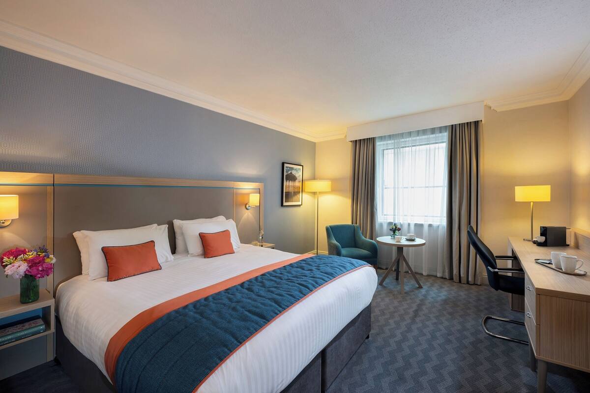 A hotel room with a large bed, a desk, a chair, a small table, and a window with curtains.