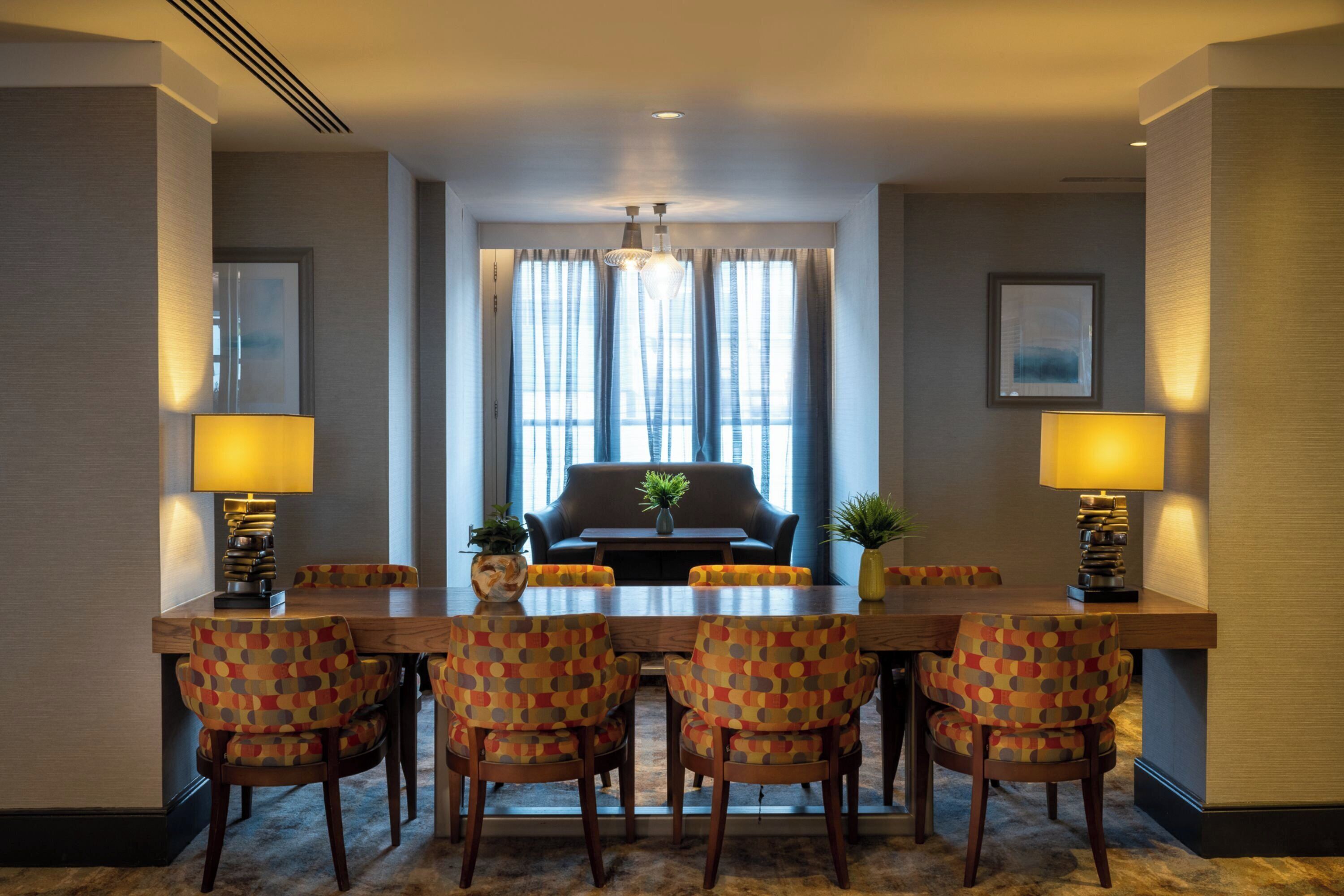 A hotel dining area with a long table, six patterned chairs, two lamps, a sofa, and a large window with curtains.
