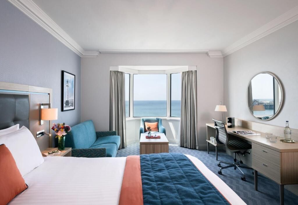 A hotel room with a bed, desk, chair, and a view of the ocean through large windows.