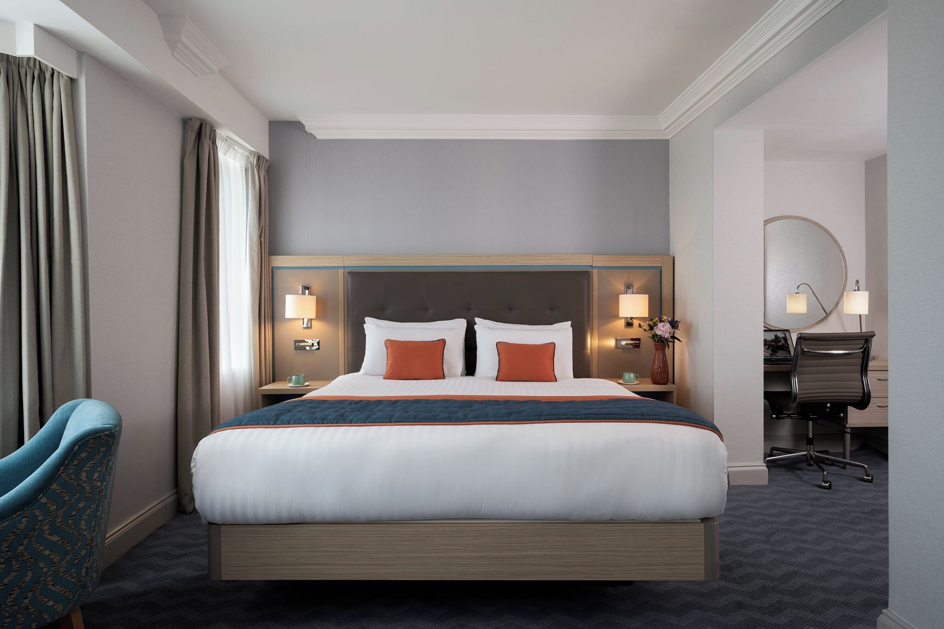 A hotel room with a large bed, two bedside tables with lamps, a desk with a chair, and a chair with a patterned cushion.