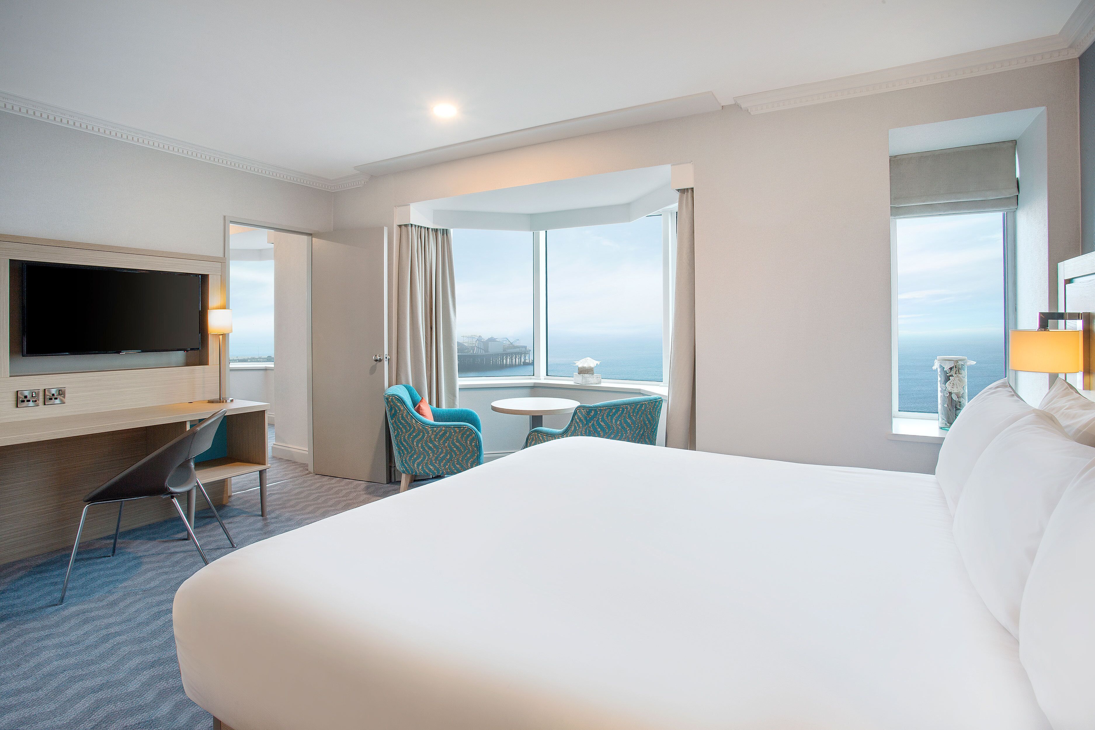 Ein Hotelzimmer mit einem großen Bett, einem an der Wand montierten Fernseher, einem Schreibtisch mit Stuhl, einem kleinen runden Tisch und Meerblick durch große Fenster.