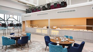 A spacious hotel dining area with multiple round tables, blue upholstered chairs, and a buffet counter with a variety of food items.