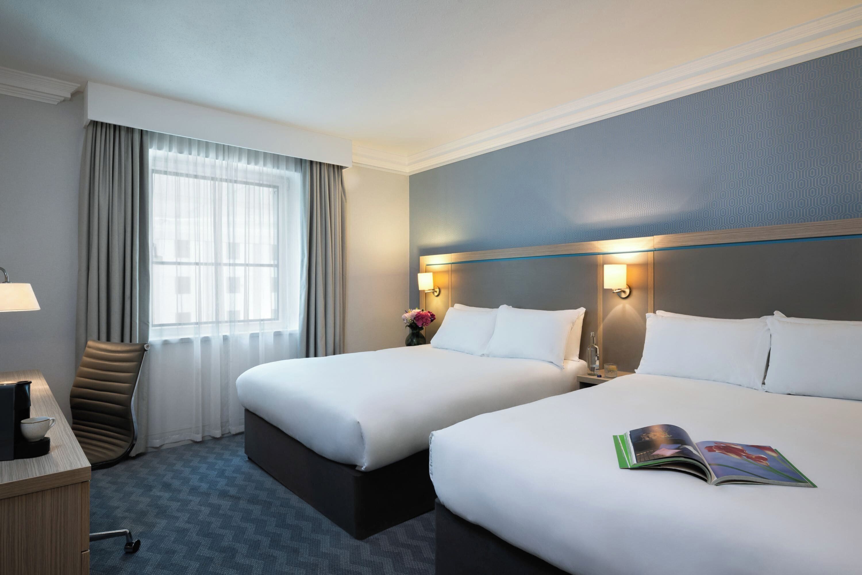 A hotel room with two beds, a desk with a lamp, a chair, and a window with curtains.