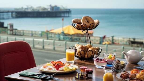 A breakfast table with croissants, fruit, and a teapot by a seaside view.