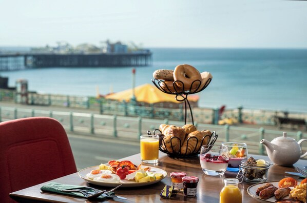 A breakfast table with croissants, fruit, and a teapot by a seaside view.