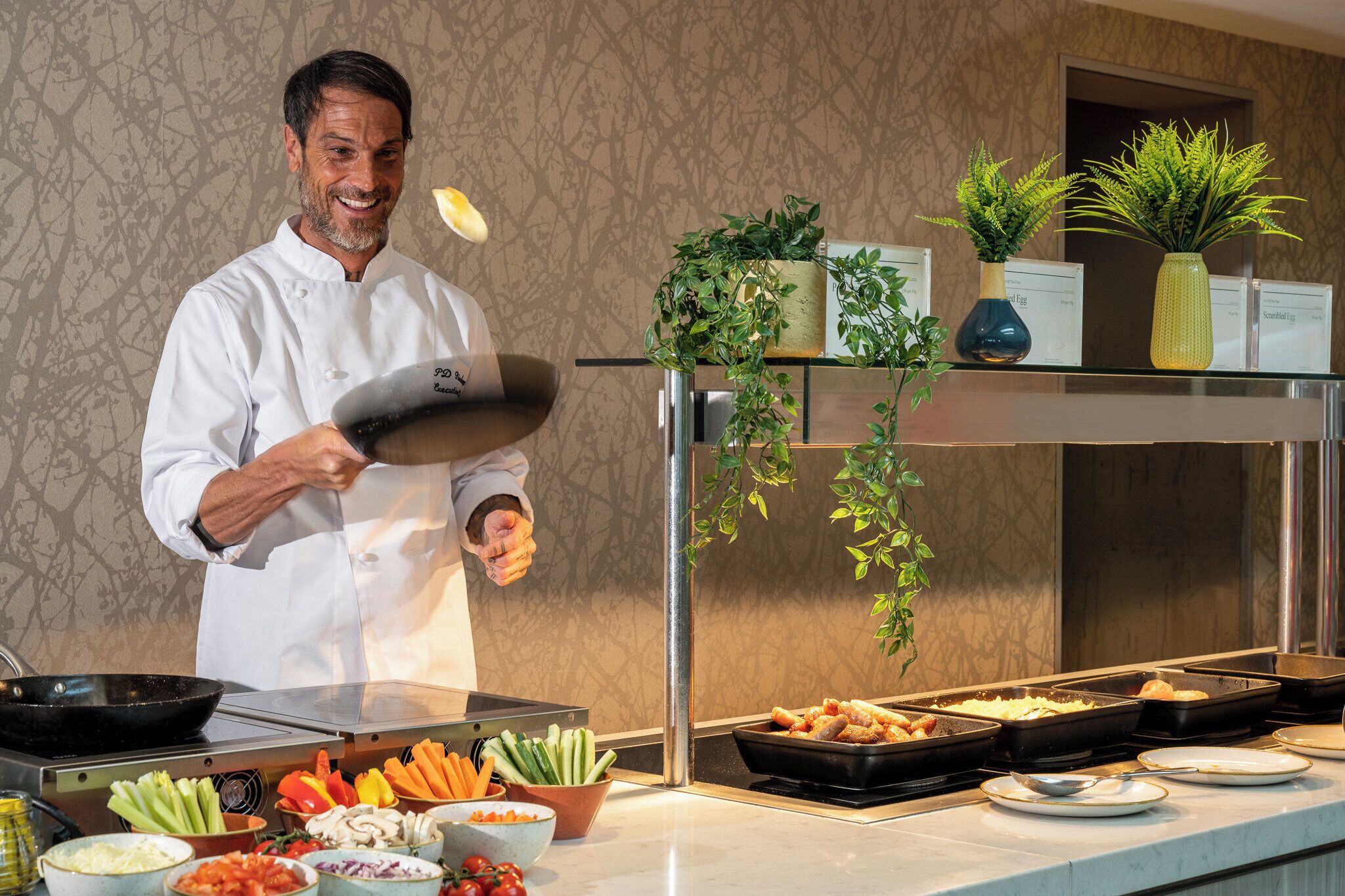 A chef in a white uniform tossing food in a kitchen with various food items and plants on display.
