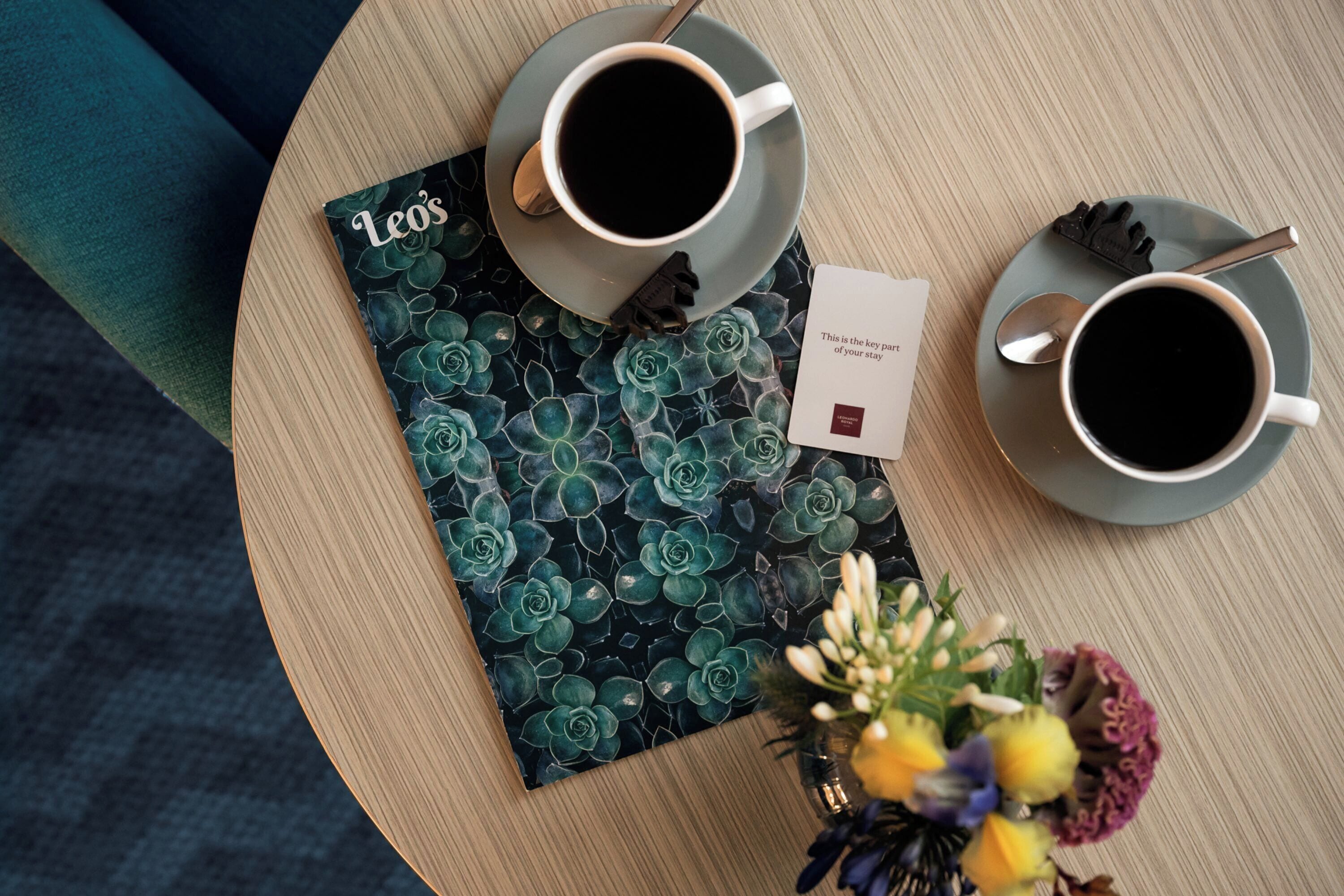 Ein Tisch mit zwei Kaffeetassen, einem Buch mit dem Titel 'Leo's', einer kleinen Karte und einer Vase mit Blumen.