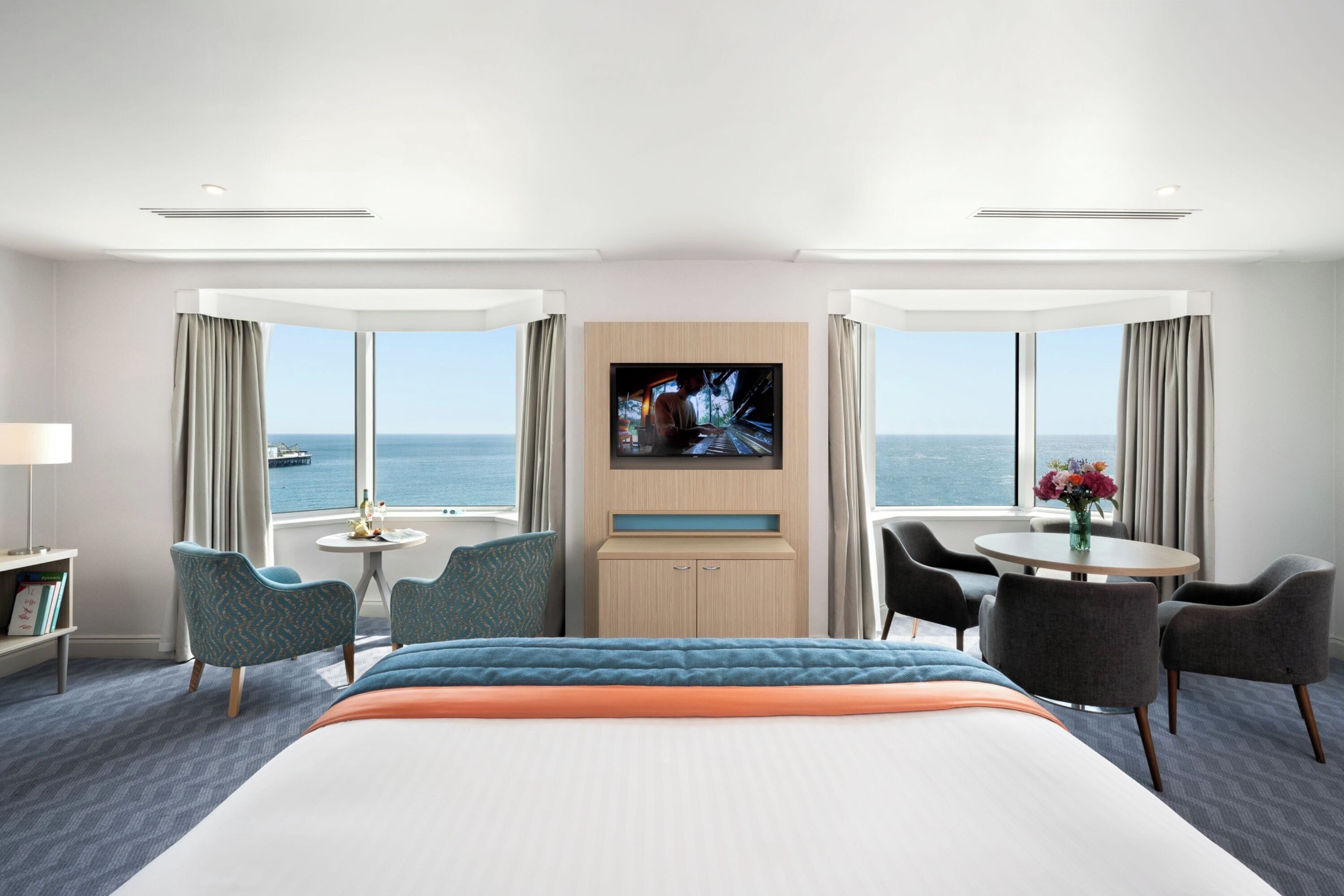 A hotel room with a bed, a TV, a desk, and a view of the sea.