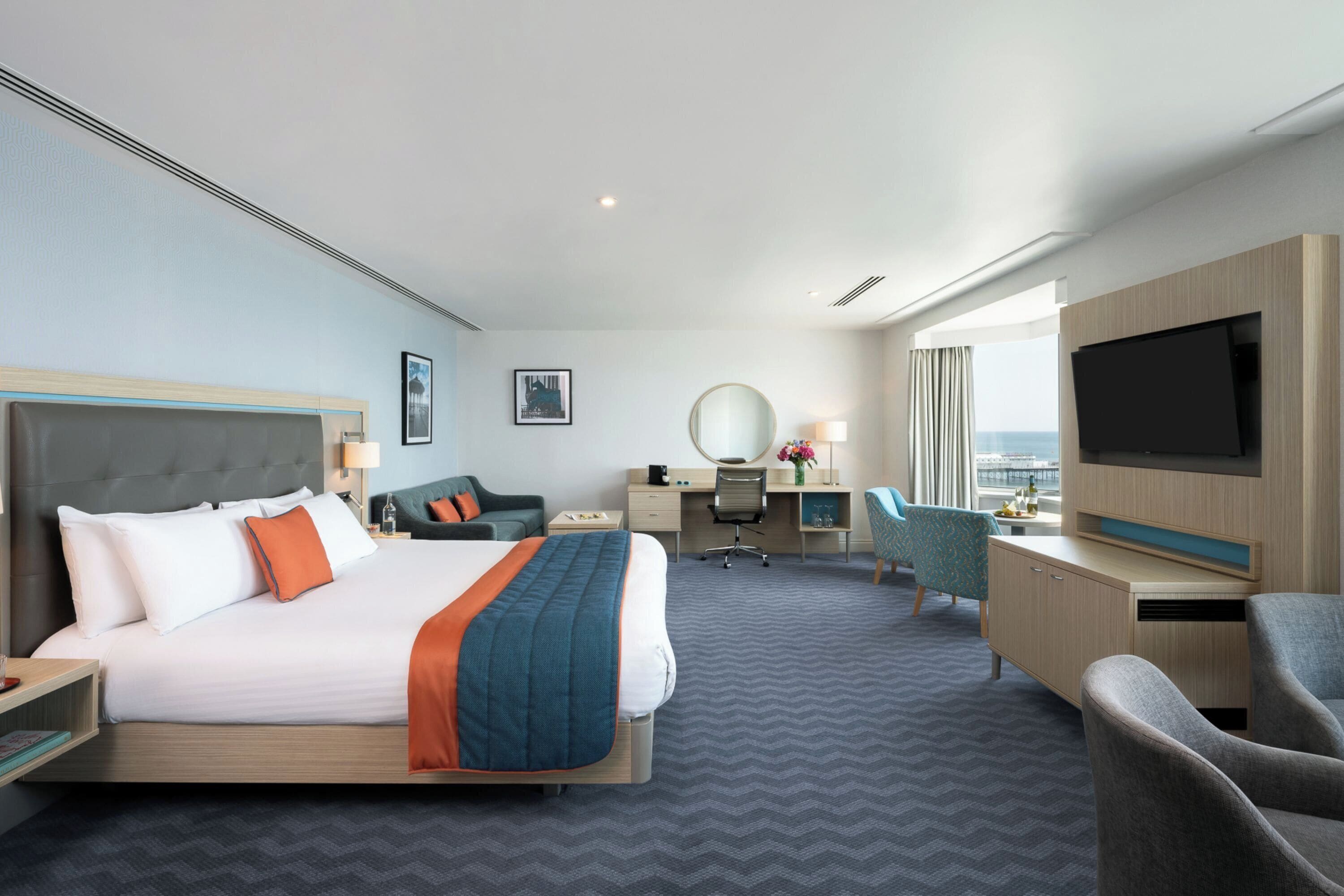 A hotel room with a large bed, a desk, a chair, a TV, and a view of the ocean.