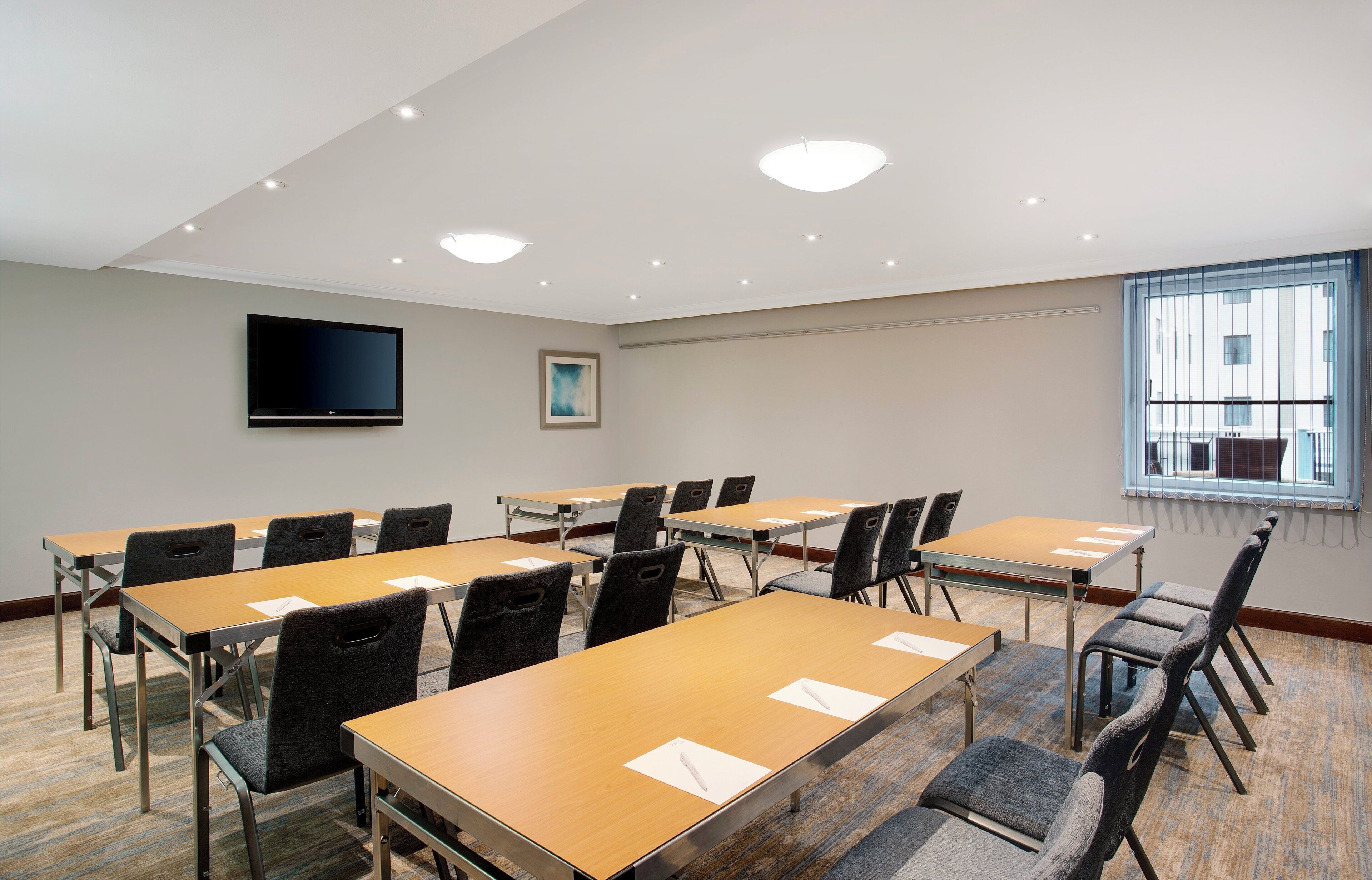 A conference room with multiple tables and chairs, a flat-screen TV mounted on the wall, and a window with blinds.