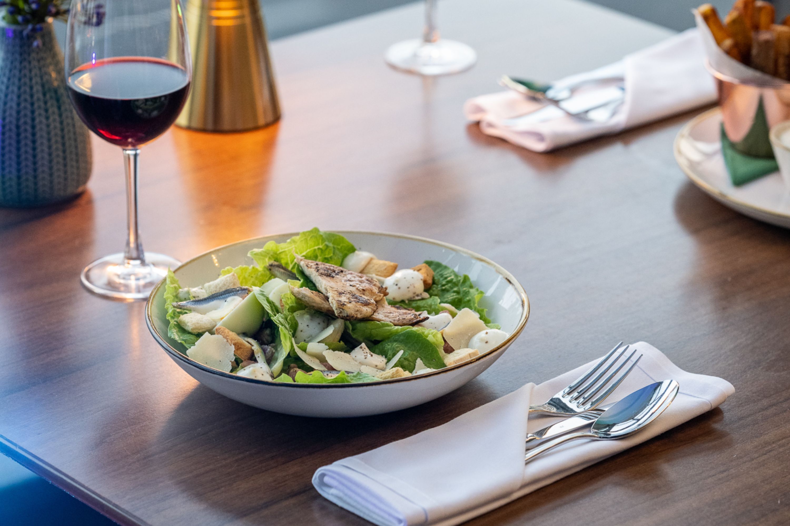 A dining table with a bowl of salad, a glass of red wine, and a folded napkin with cutlery.