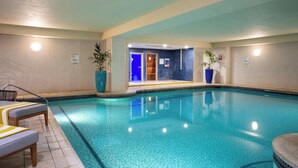 An indoor swimming pool with clear blue water, surrounded by tiled walls and a ceiling with recessed lighting. There are lounge chairs and potted plants along the poolside.
