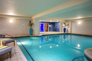 An indoor swimming pool with clear blue water, surrounded by tiled walls and a ceiling with recessed lighting. There are lounge chairs and potted plants along the poolside.
