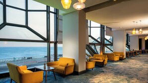A spacious lobby with large windows offering a view of the sea, yellow armchairs, and a modern staircase.
