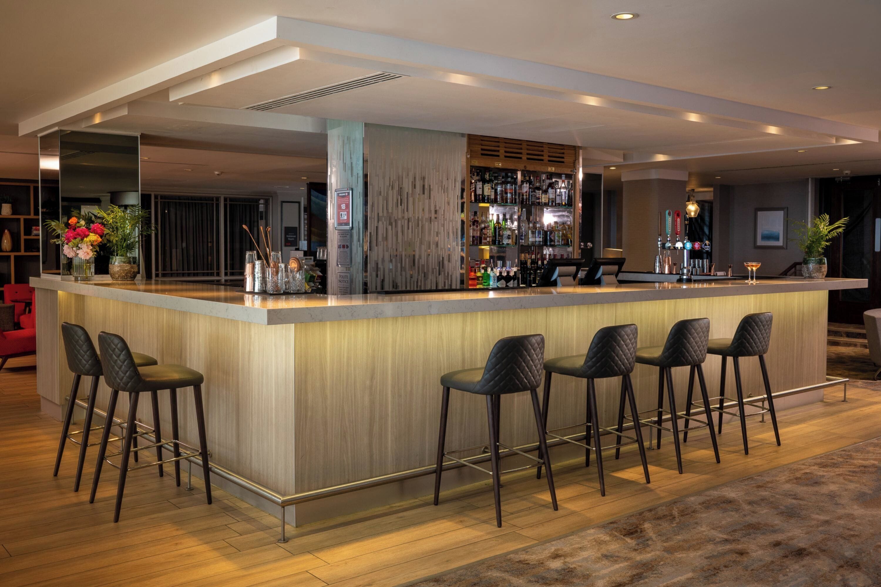 A modern bar area with a wooden counter, black bar stools, and a variety of bottles and glasses.