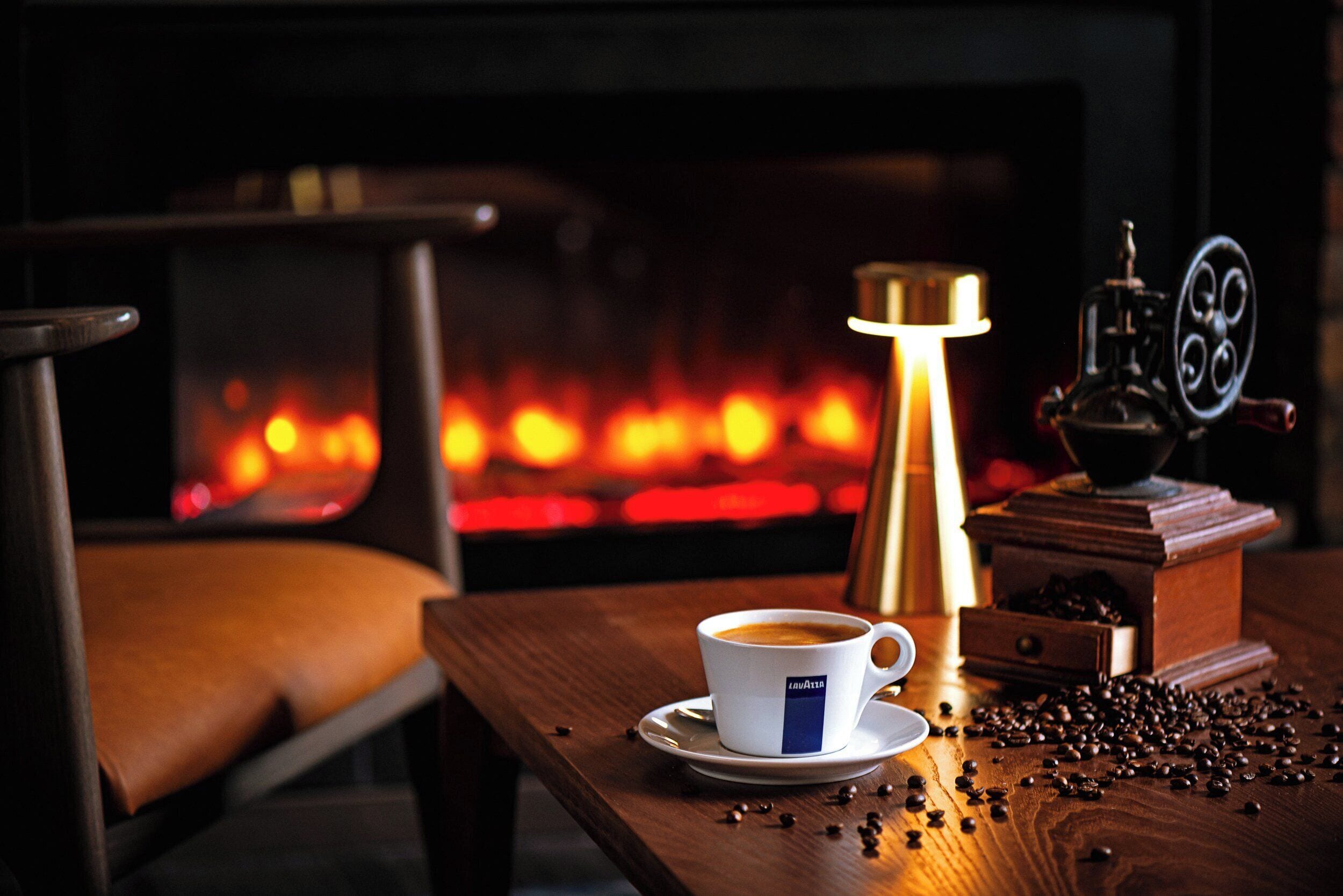 Una taza de café sobre un plato, junto a un molinillo de café y granos de café esparcidos sobre una mesa de madera.
