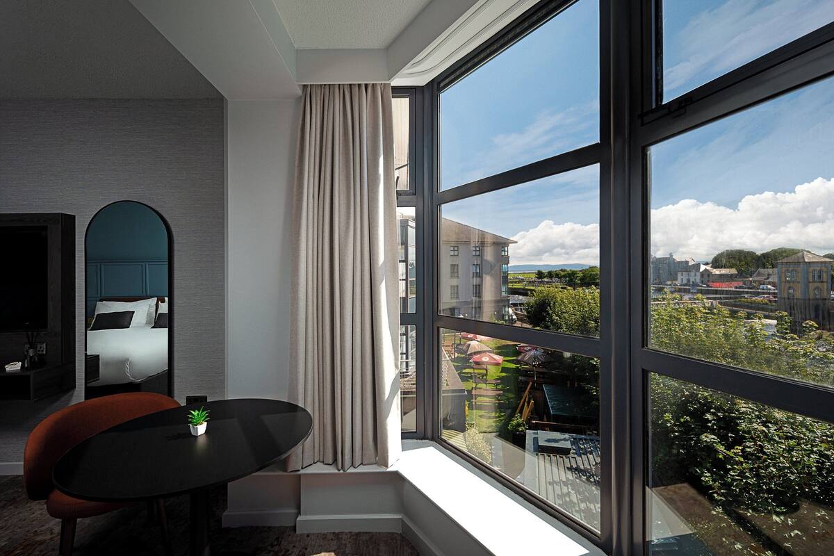 A modern hotel room with a large window, a small table with a potted plant, a chair, and a bed with a mirror.