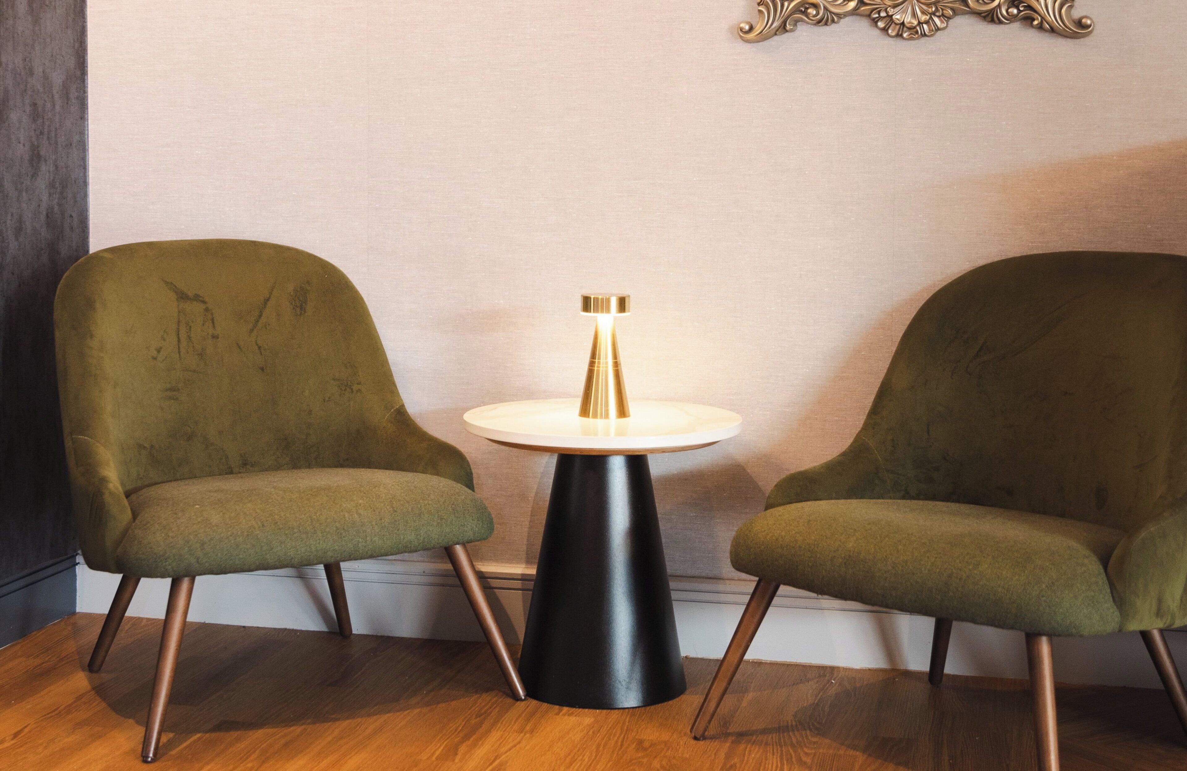 Twee groene fauteuils en een kleine ronde tafel met een zwart onderstel en een gouden lamp in een kamer met houten vloeren en een effen muur.