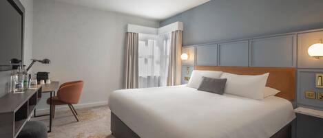 Habitación de hotel con una cama grande, un escritorio con silla, un espejo y una ventana con cortinas.