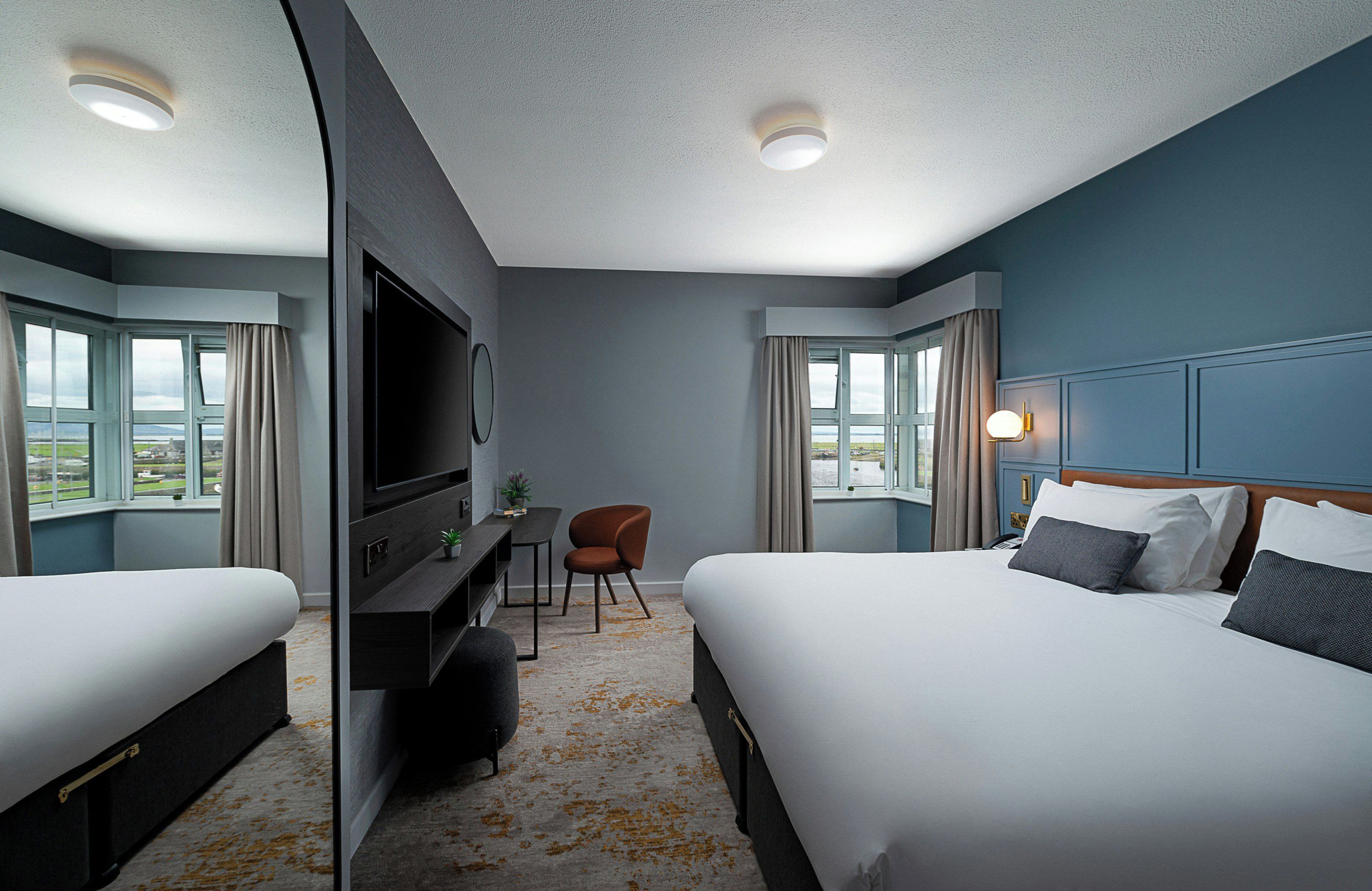 A hotel room with two beds, a desk, a chair, a television, and a view of the outdoors.