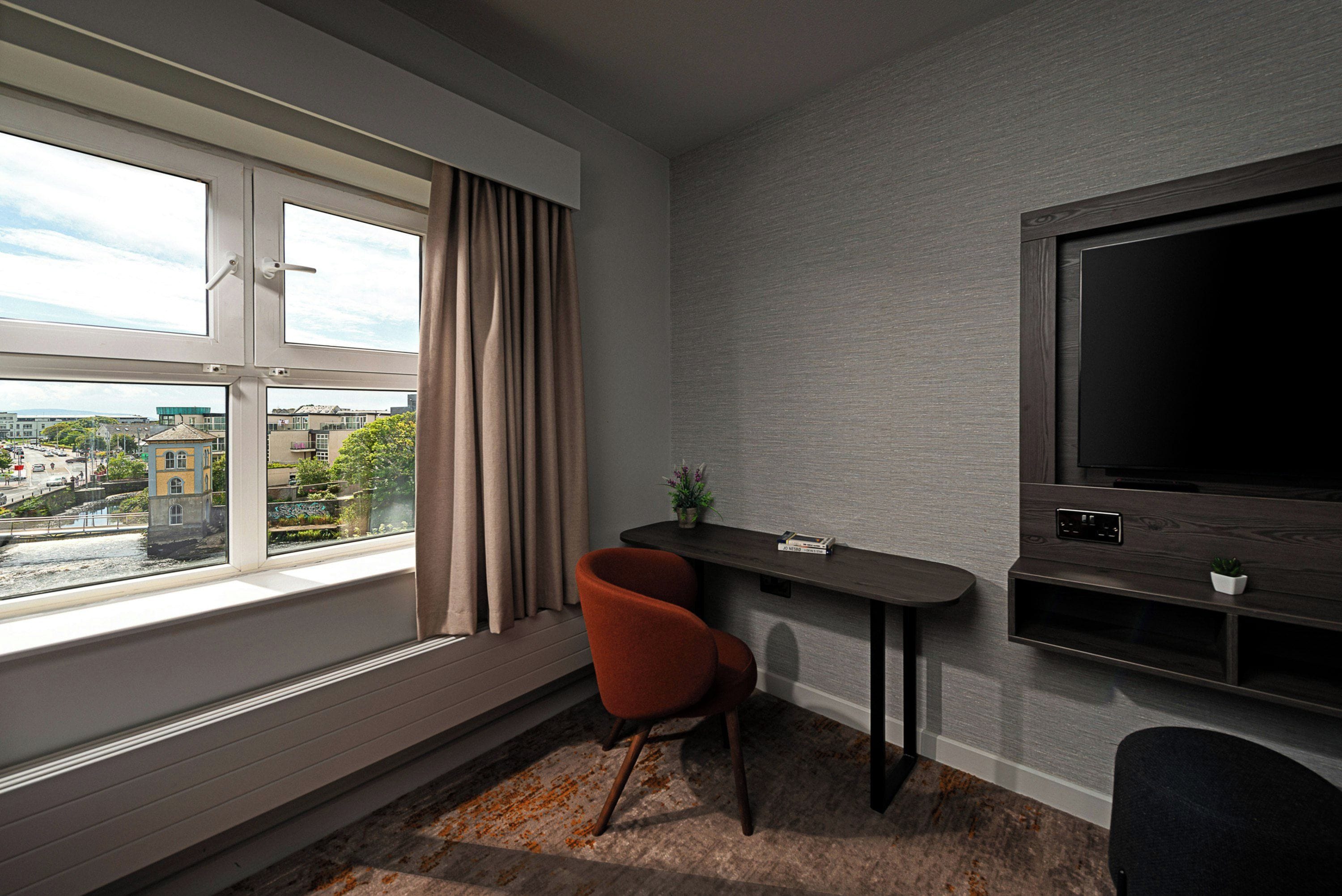 Een moderne kamer met een groot raam, een flatscreen televisie, een klein tafeltje en een rode stoel.
