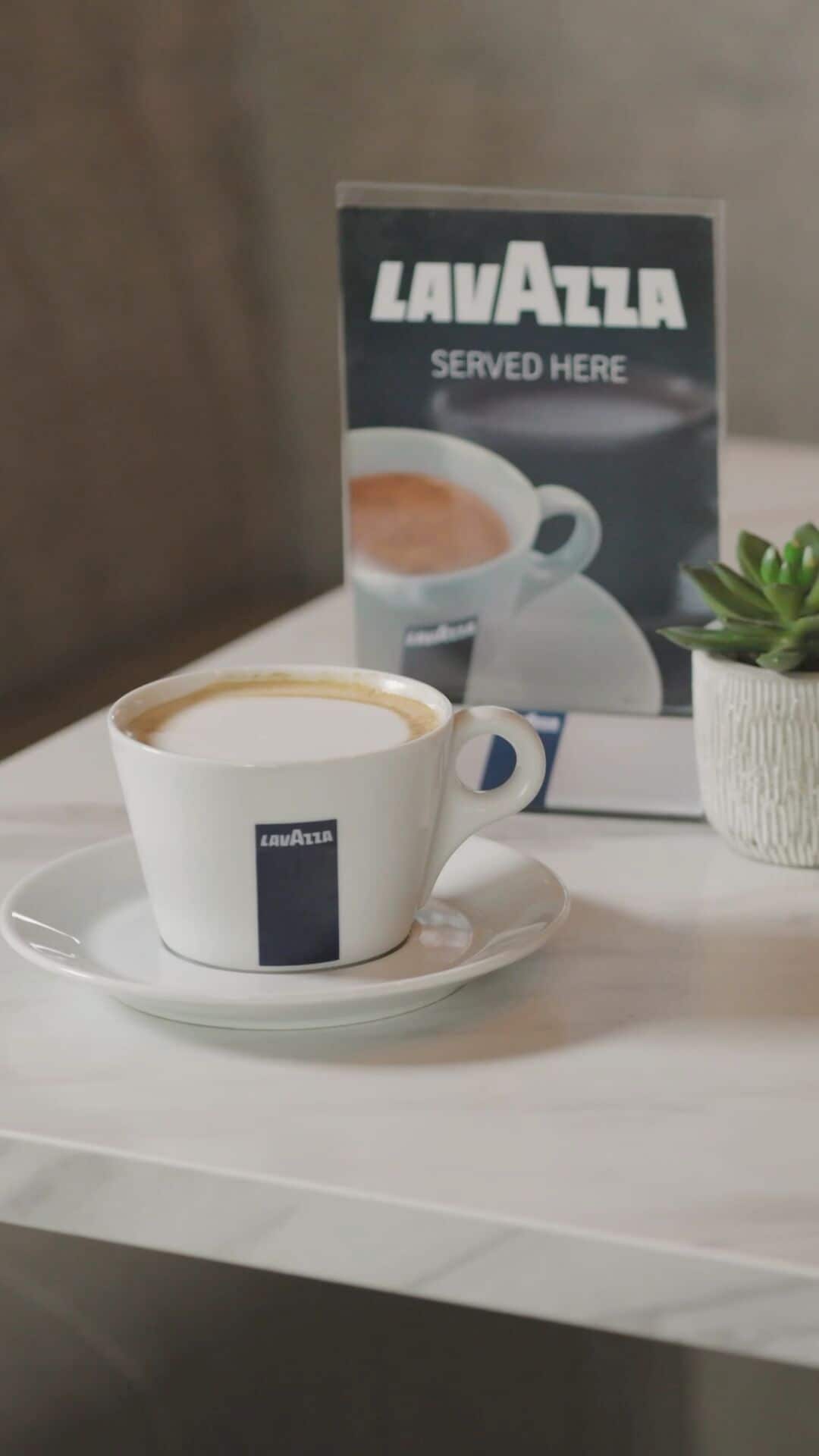 Una taza de café Lavazza sobre un platillo, con un cartel anunciando el café al fondo.