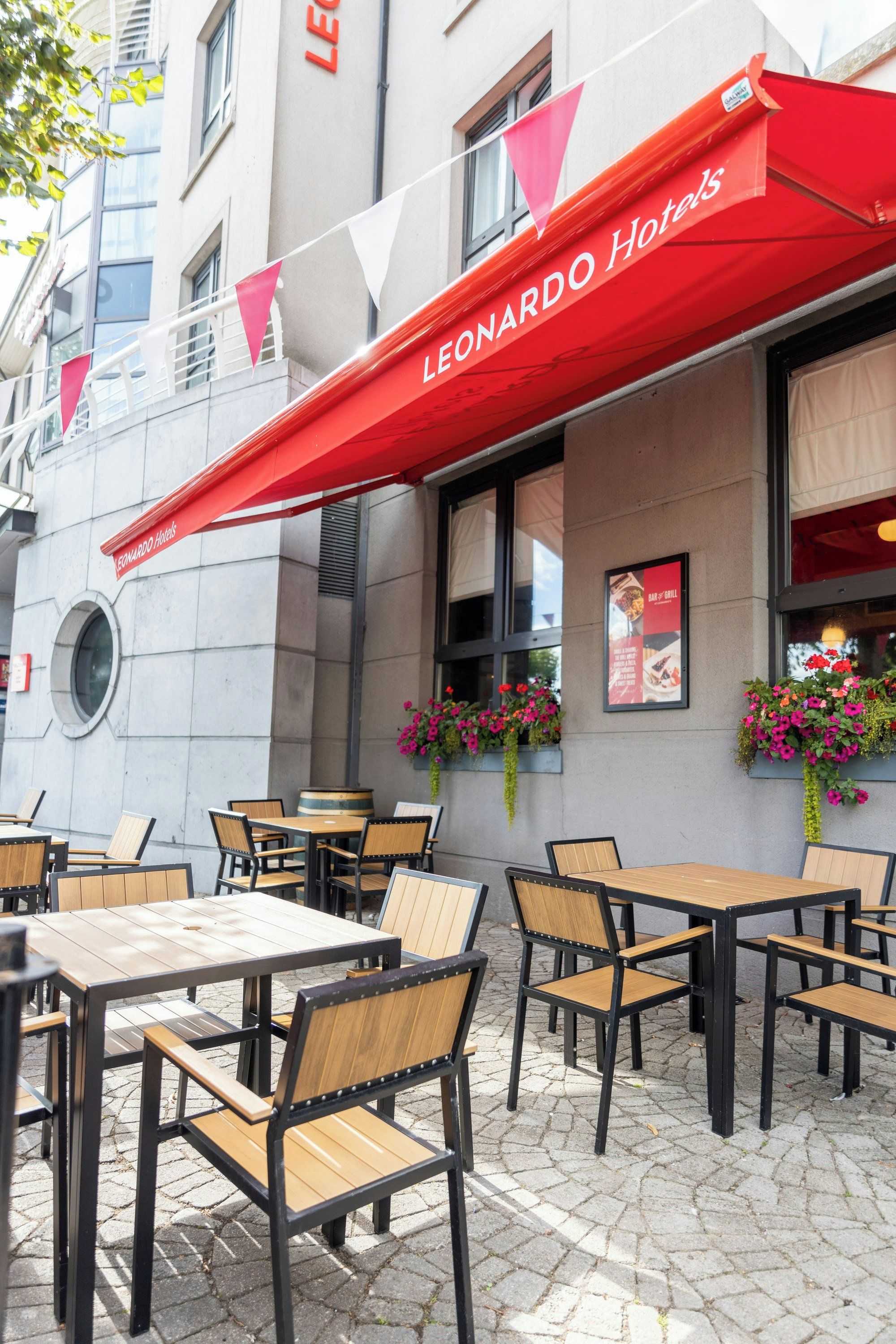 Mobiliario exterior con mesas y sillas, un toldo rojo con la marca 'LEONARDO Hotels' y macetas con flores en la fachada del edificio.