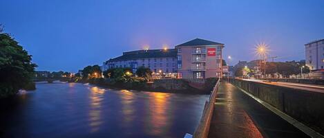 A hotel with multiple floors and balconies, situated beside a river with a bridge in the background.