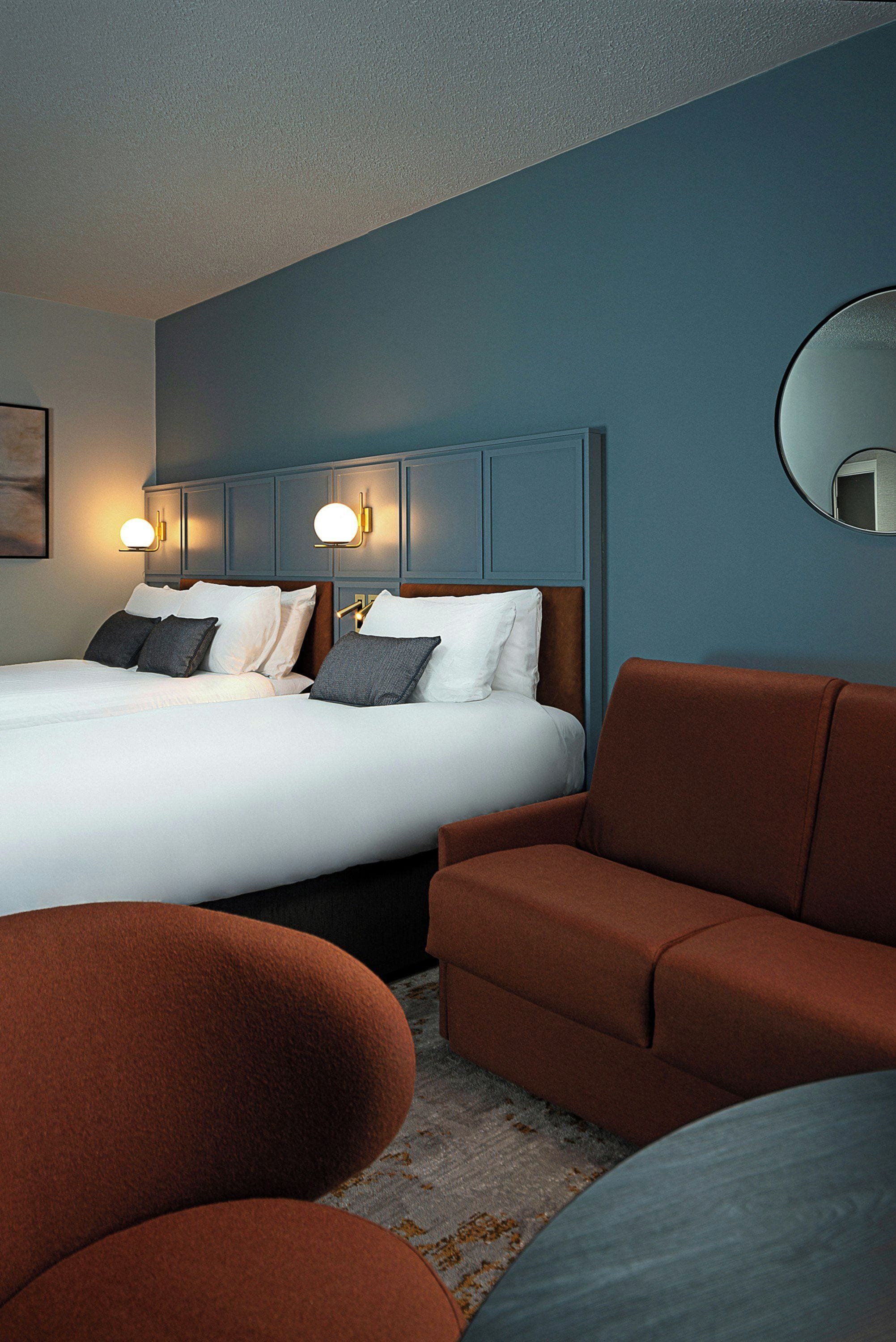 Habitación de hotel con una cama, dos mesitas de noche, un espejo redondo y una zona de estar con dos sofás.