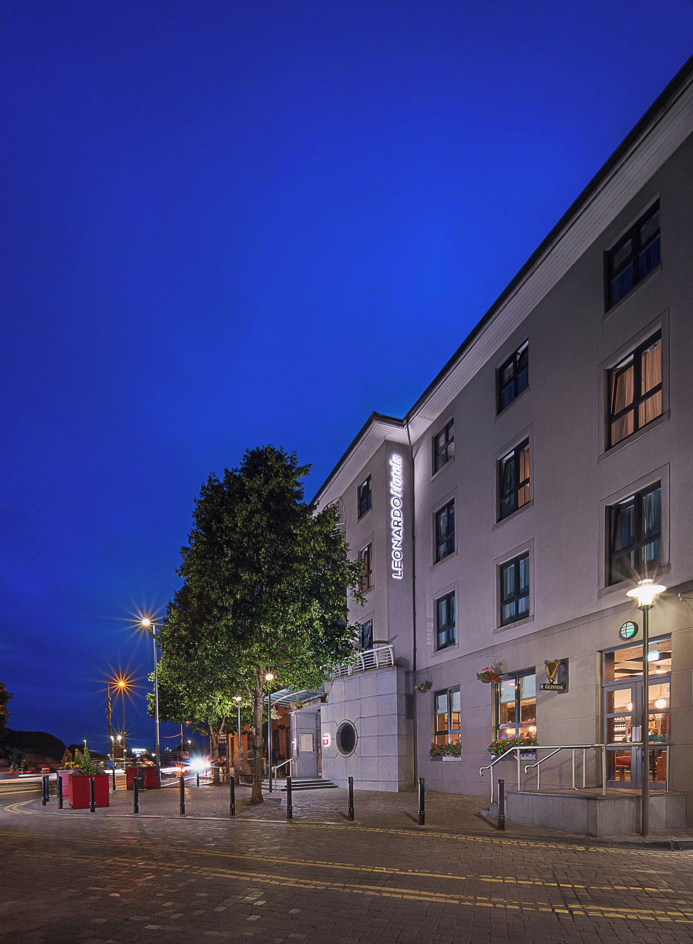 Ein Hotelgebäude mit klarem Himmel im Abendrot, ein Baum im Vordergrund und eine Straße mit Absperrbügeln und einem roten Container.