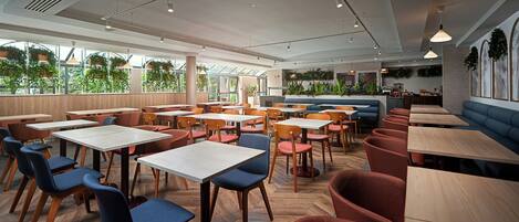 A modern restaurant interior with multiple seating options including tables and booths, large windows with greenery outside, and a variety of pendant lighting fixtures.