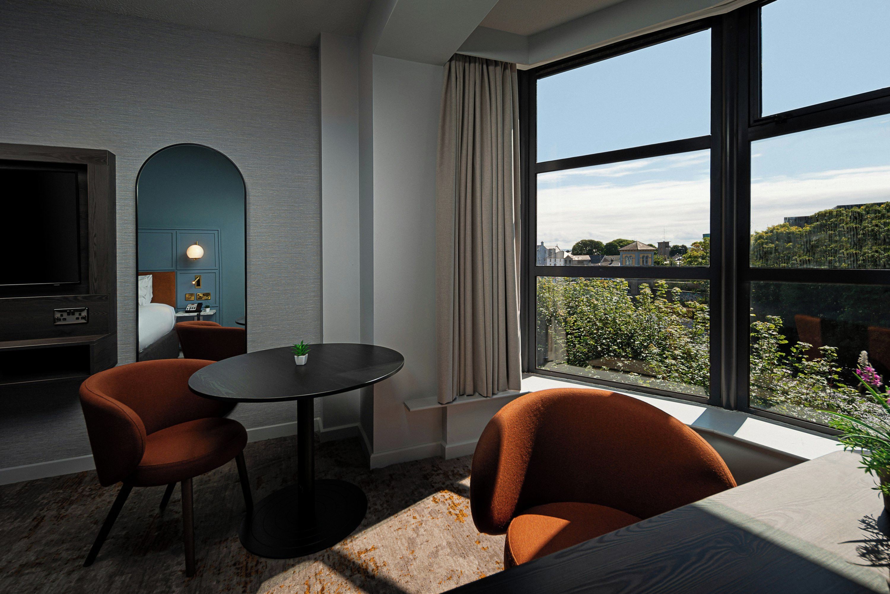 A modern hotel room with a large window, a flat-screen TV, two orange chairs, a small table, and a plant.