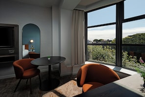 Habitación de hotel moderna con un ventanal grande, un televisor de pantalla plana, dos sillones naranjas, una mesita y una planta.