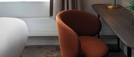 A modern hotel room with a large window, a wooden desk, a brown chair, and a white bed.
