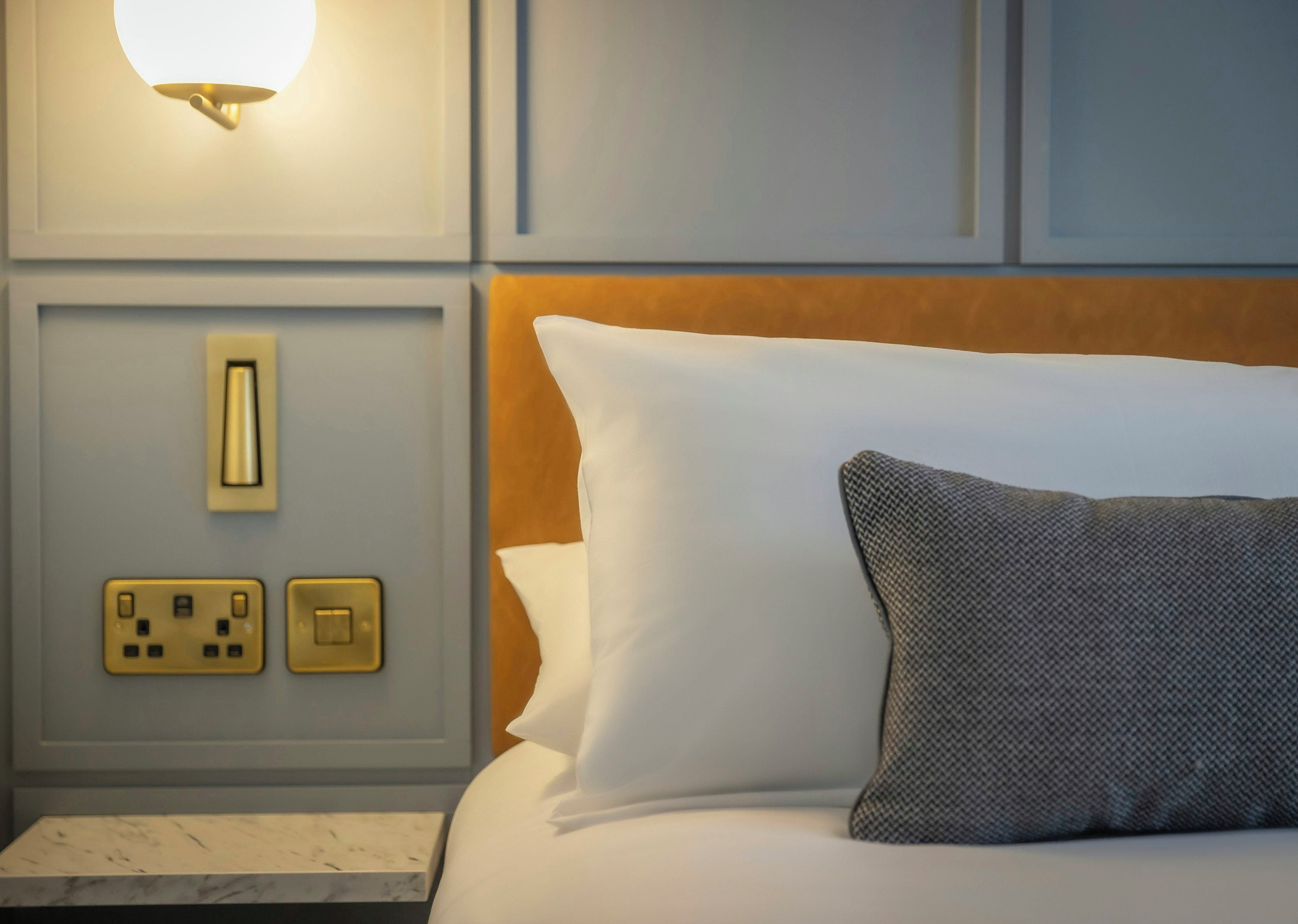 Una habitación de hotel con cama, mesita de noche, luz empotrada en la pared y enchufes.