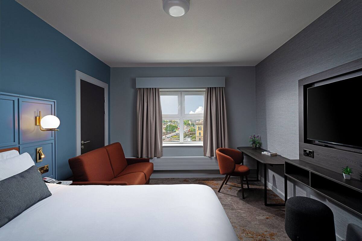A hotel room with a large bed, a desk, a TV, and a view of the city through the window.