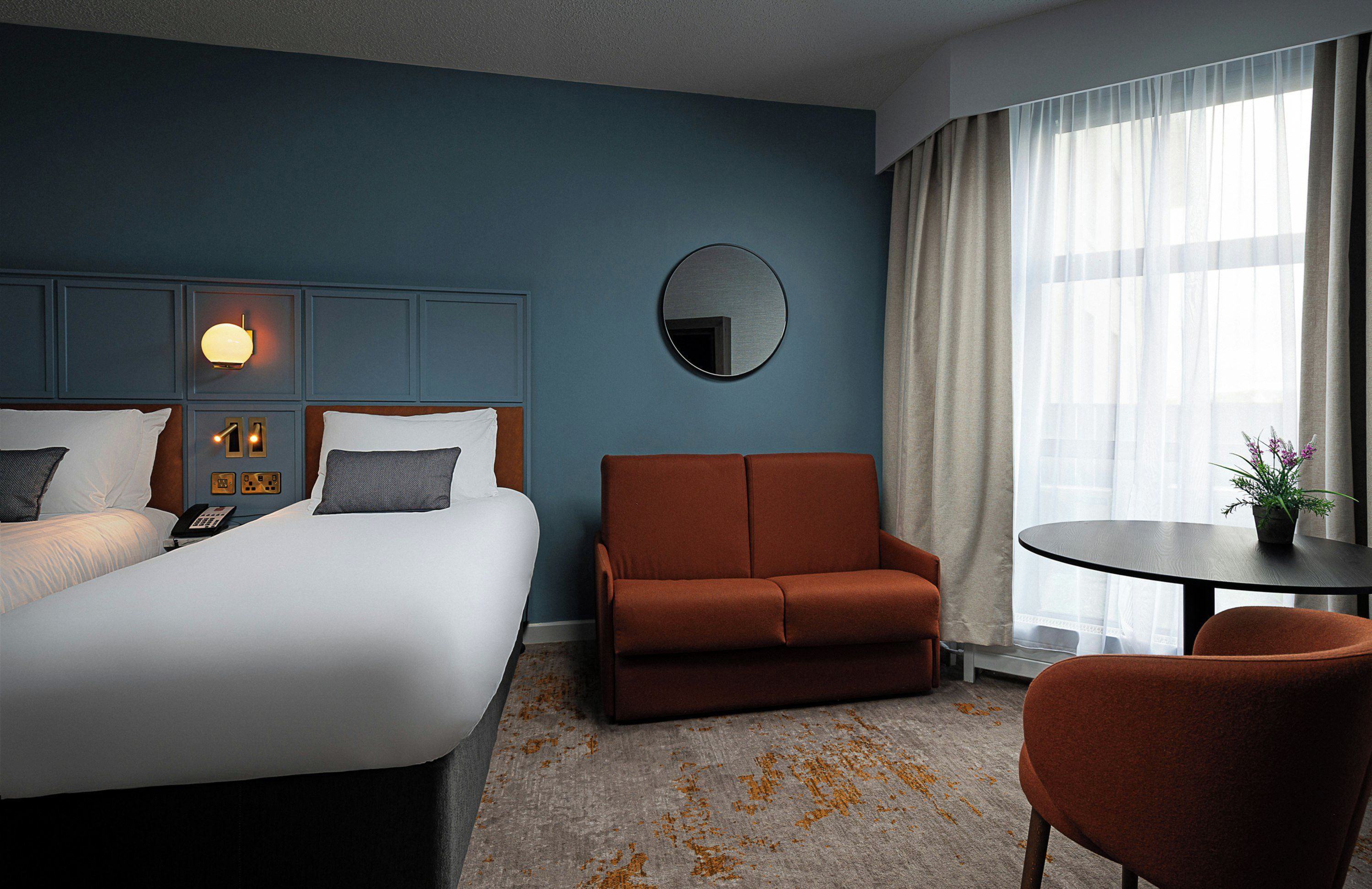 A hotel room with a bed, a sofa, a chair, a small table, a mirror, and a window with curtains.