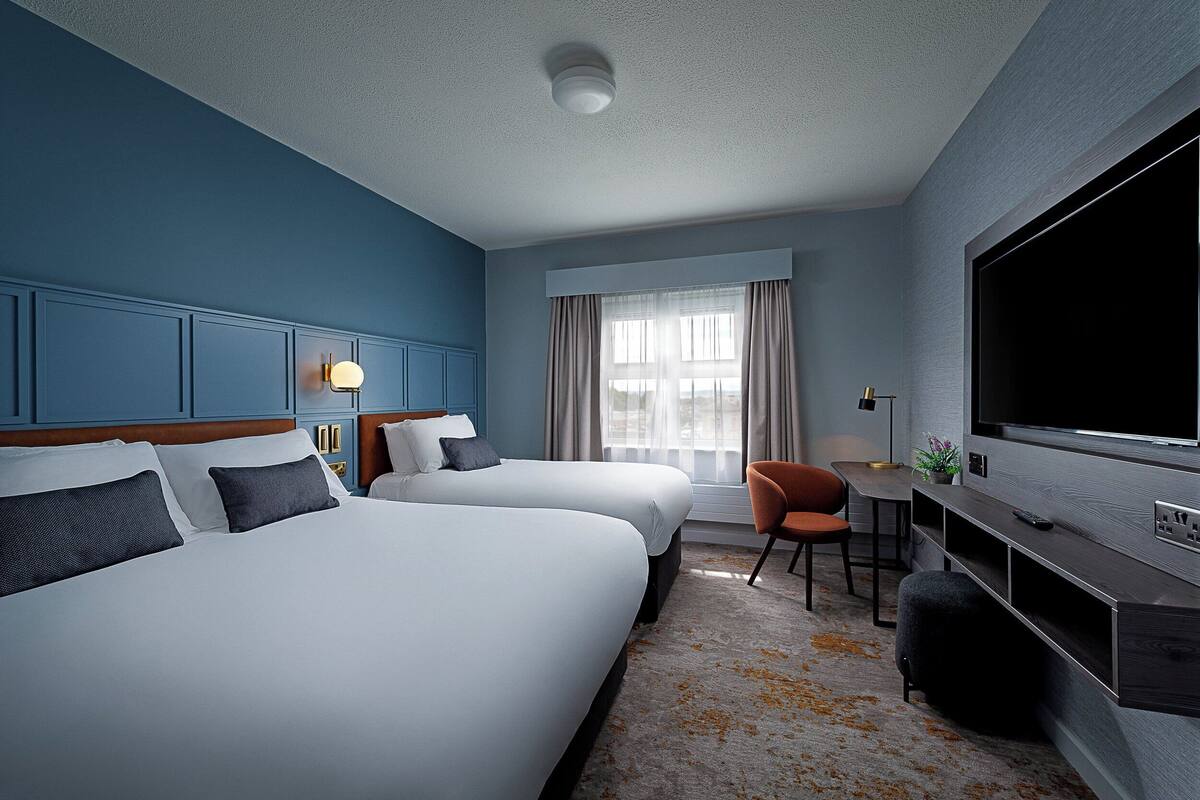A hotel room with two beds, a TV, a desk, and a window with curtains.