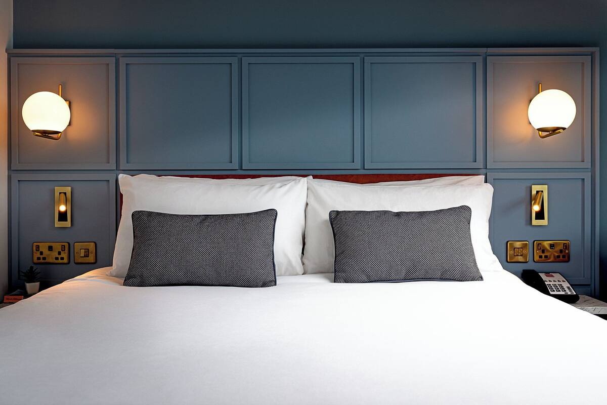A neatly made bed with white and patterned pillows, a wooden headboard, and wall-mounted lighting fixtures.