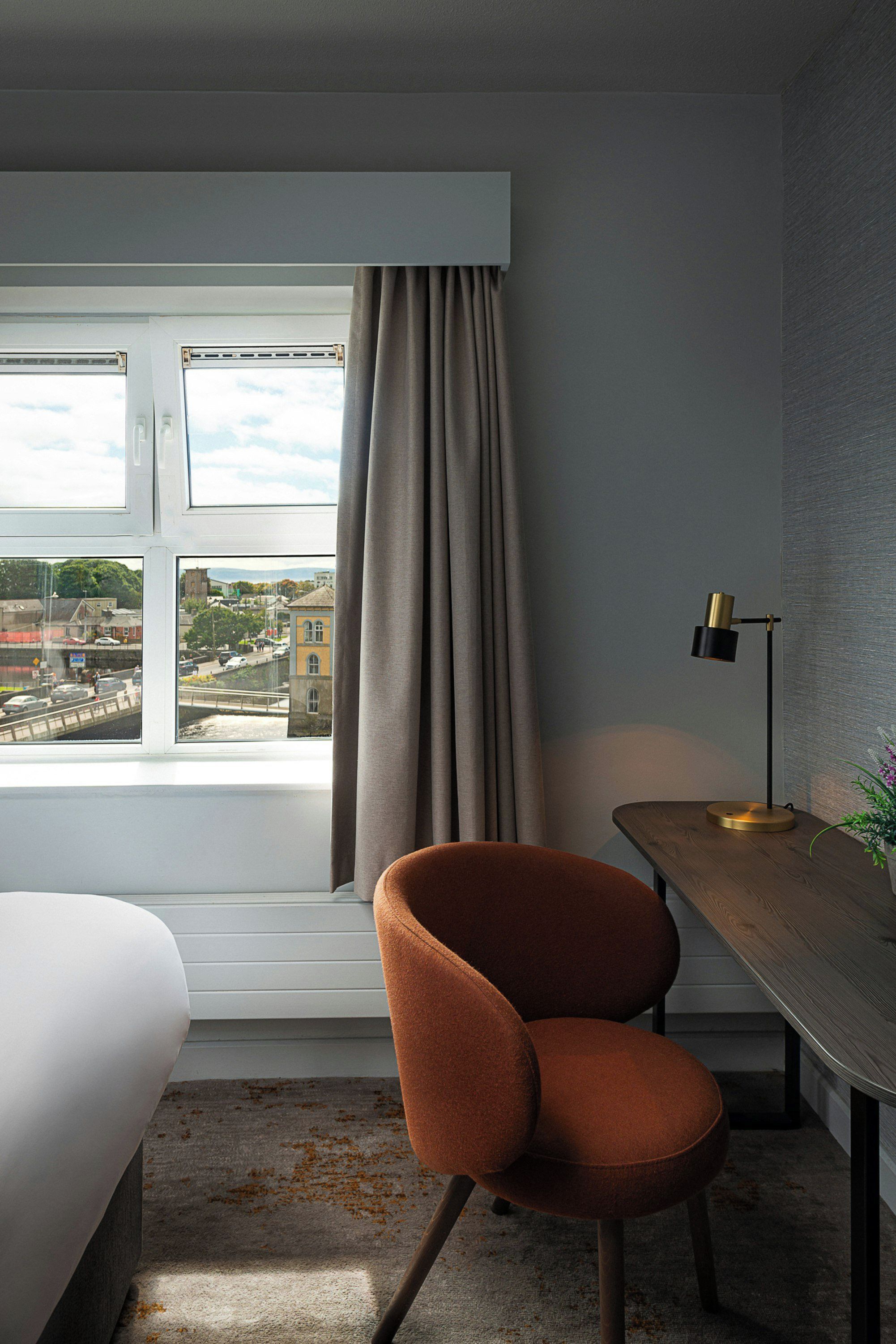 A hotel room with a bed, a desk, a chair, a window with curtains, and a view of the cityscape.