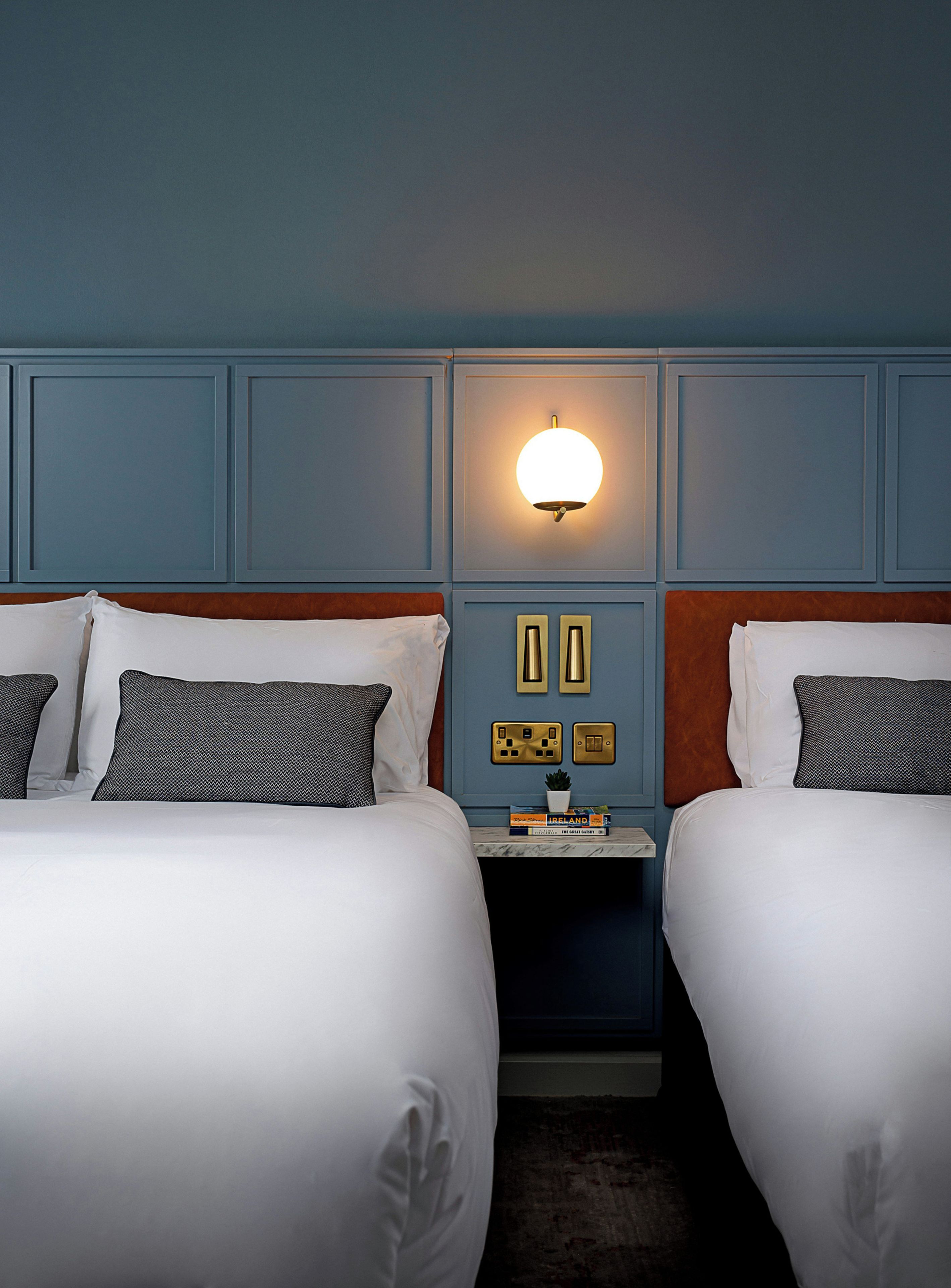 Una camera d'albergo con due letti, un comodino con una lampada e un punto luce a parete.